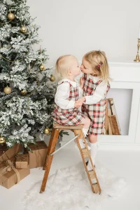 Julfotografering i fotostudio med två barn som pussas framför en pyntad julgran. 