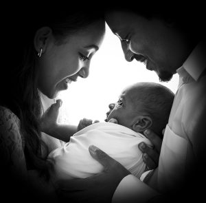 Svartvitt familjeporträtt där mamma och pappa ler mot den nyfödda bebisen i deras famn. 
